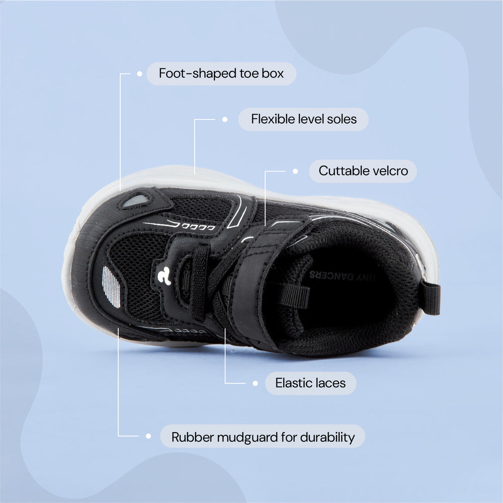 Play Infant in black from above, labelled: foot-shaped toe box, flexible level soles, cuttable velcro, elastic laces, rubber mudguard.