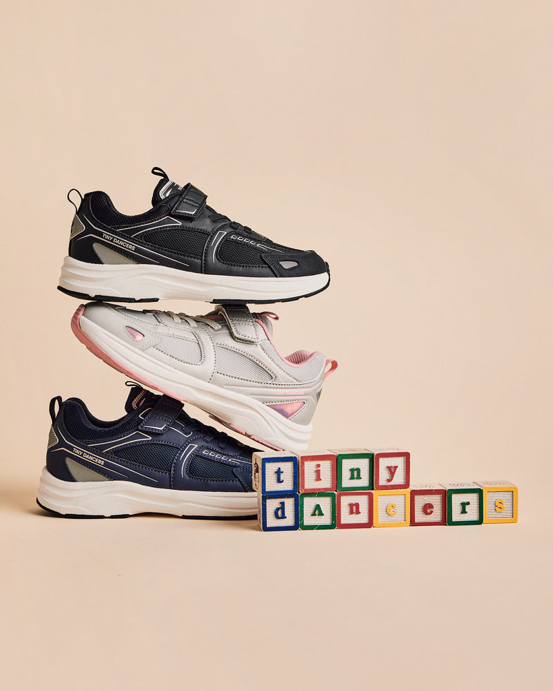 Three pairs of Play Junior shoes stacked. Black on top, Silver/Pink in the middle, Navy on the bottom, with wooden alphabet blocks spelling "tiny dancers" alongside.