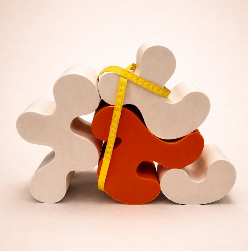 A yellow measuring tape wrapped around sculptural wooden shapes on a soft pink background.